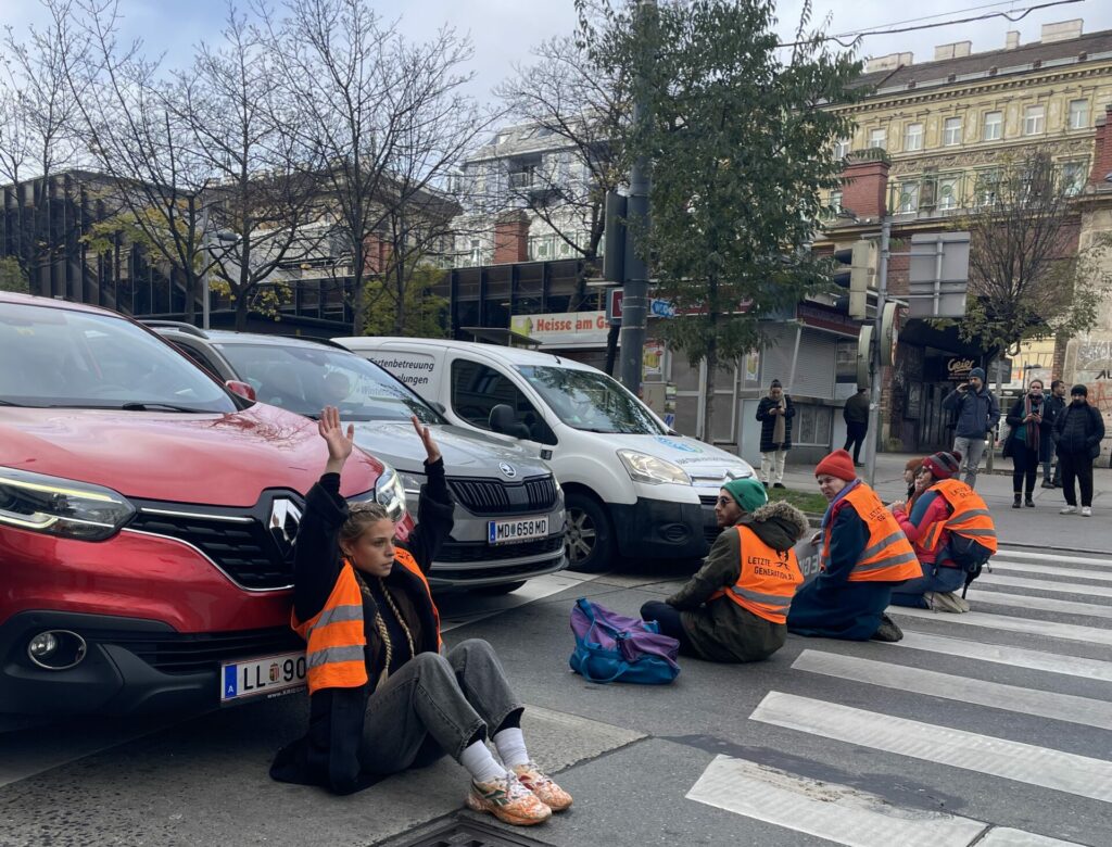 Aktivist:innen der Letzten Generatin sitzen auf einem Zebrastreifen vor drei Autos.