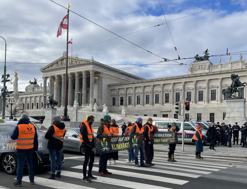 Aktivist:innen der Letzten Generation blockieren einen Zebtrastreifen vor dem Parlament in Wien. Hinten im Bild stehen Polizist:innen und Polizeiwägen.